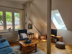 A cozy room with a sloped ceiling, a sofa, a coffee table, a bookshelf, and a window with a view of greenery. - Wustrow Parish Barn (Wustrow)
