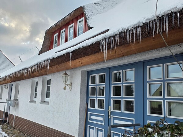 Un bâtiment à deux étages, avec un toit rouge, des murs blancs et des portes bleues. Des glaçons pendent du toit, et on peut voir une fondation en briques.