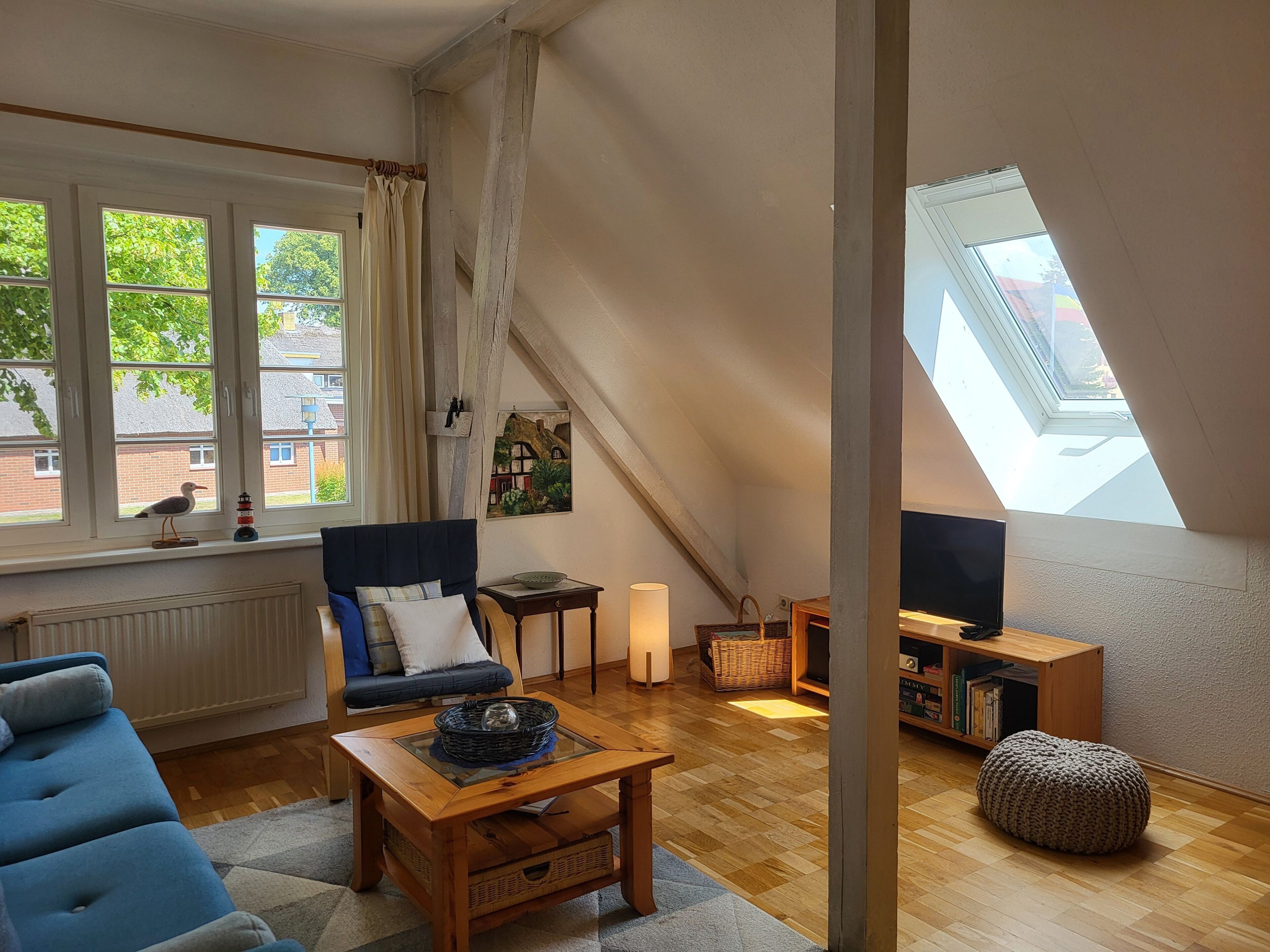 A cozy room with a sloped ceiling, a sofa, a coffee table, a bookshelf, and a window with a view of greenery.