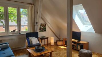 A cozy room with a sloped ceiling, a sofa, a coffee table, a bookshelf, and a window with a view of greenery.