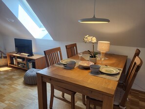 A dining area with a wooden table, chairs, a television on a stand, a plant, and a lamp.