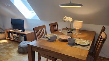 Een eethoek met een houten tafel, stoelen, een televisie op een standaard, een plant en een lamp.