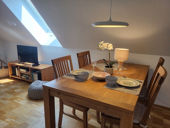 A dining area with a wooden table, chairs, a television on a stand, a plant, and a lamp.