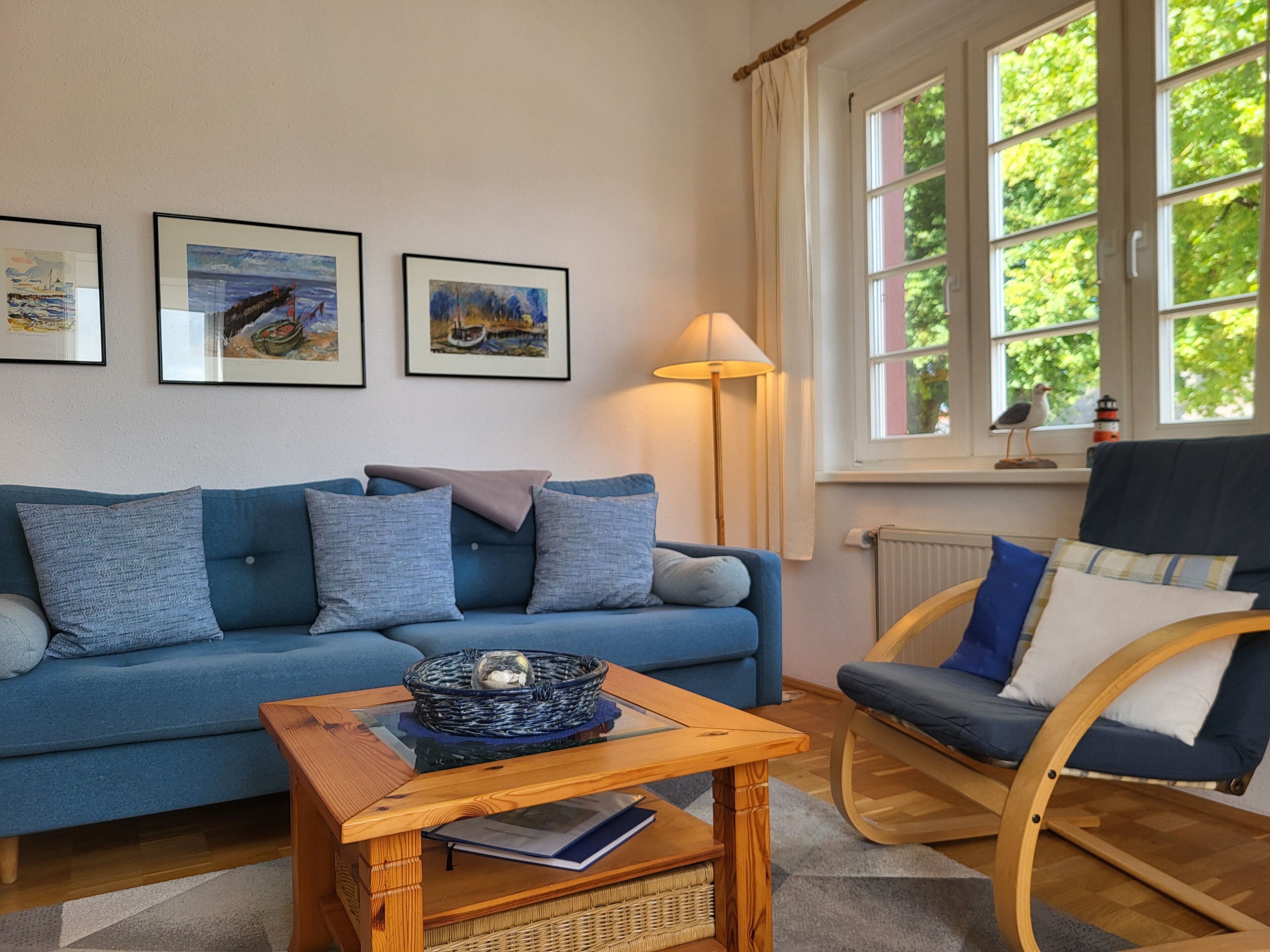Un salon avec un canapé bleu, une table basse en bois, un panier en osier et un fauteuil. Trois œuvres d’art encadrées ornent le mur.
