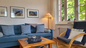 Een woonkamer met een blauwe bank, een houten bijzettafeltje, een rieten mand en een stoel. Er hangen drie kunstwerken in lijsten aan de muur.