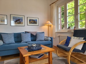 A living room with a blue sofa, a wooden coffee table, a wicker basket, and a chair. There are three framed artworks on the wall.