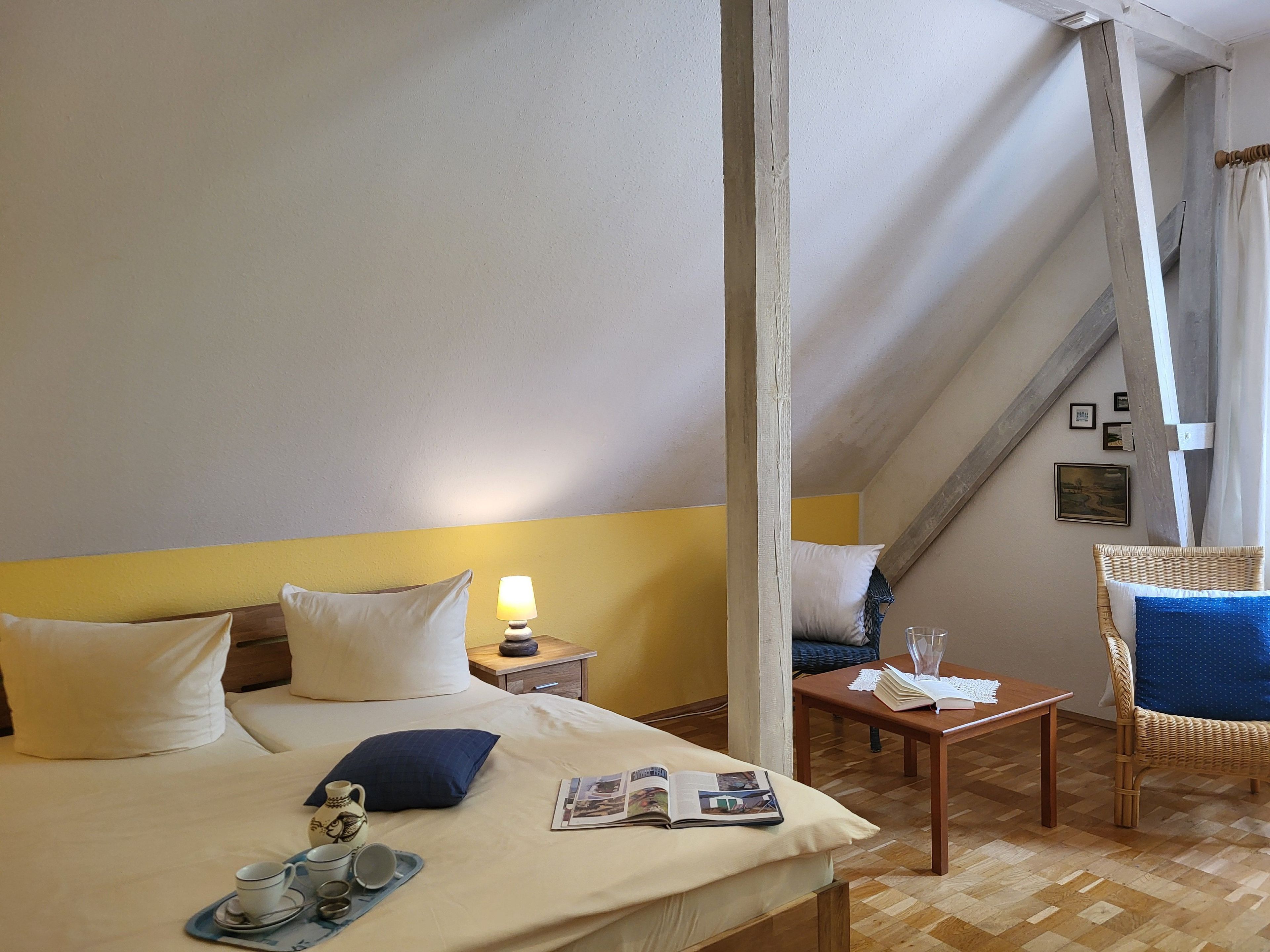 Hotelkamer met een bed, nachtkastje, lamp, stoel en een kleintje tafeltje met een boek en een glas.