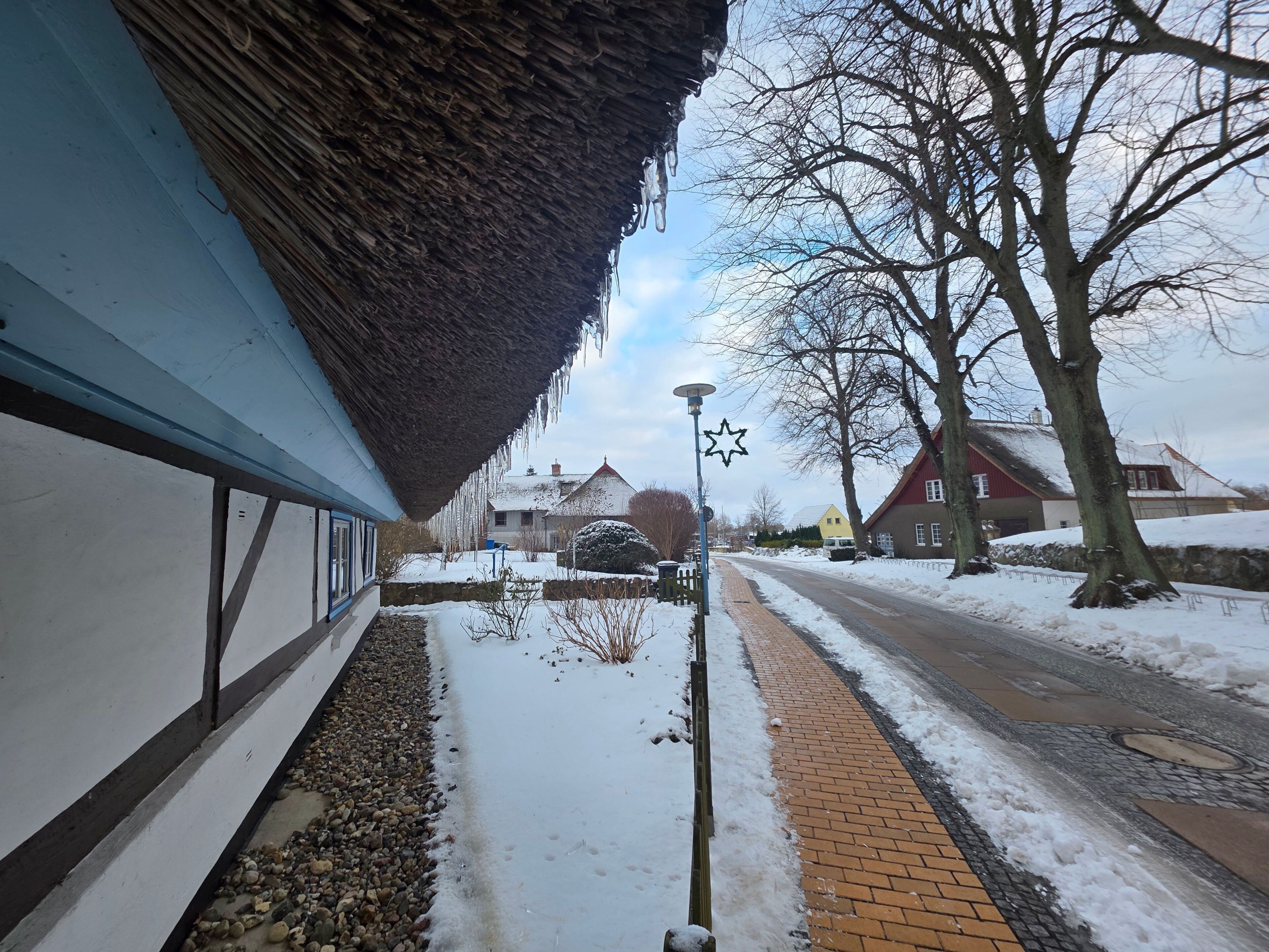 Une rue enneigée, bordée d’immeubles, avec un toit de chaume et des arbres sans feuilles.