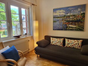 A cozy living room with a dark sofa, patterned cushions, a wicker chair, a window with a sailboat model, and a painting of a harbor scene.