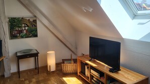 Avokeittiötyylinen huone, jossa on kalteva katto, puinen jalusta, jonka päällä on litteänäyttöinen televisio, lattiavalaisin, rottinkikori ja seinällä oleva kehystetty taulu.