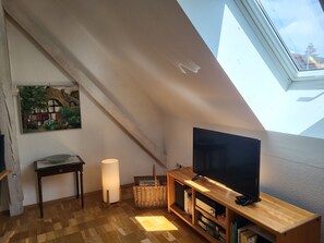A loft-style room with a sloped ceiling, a flat-screen TV on a wooden stand, a floor lamp, a wicker basket, and a framed painting on the wall.
