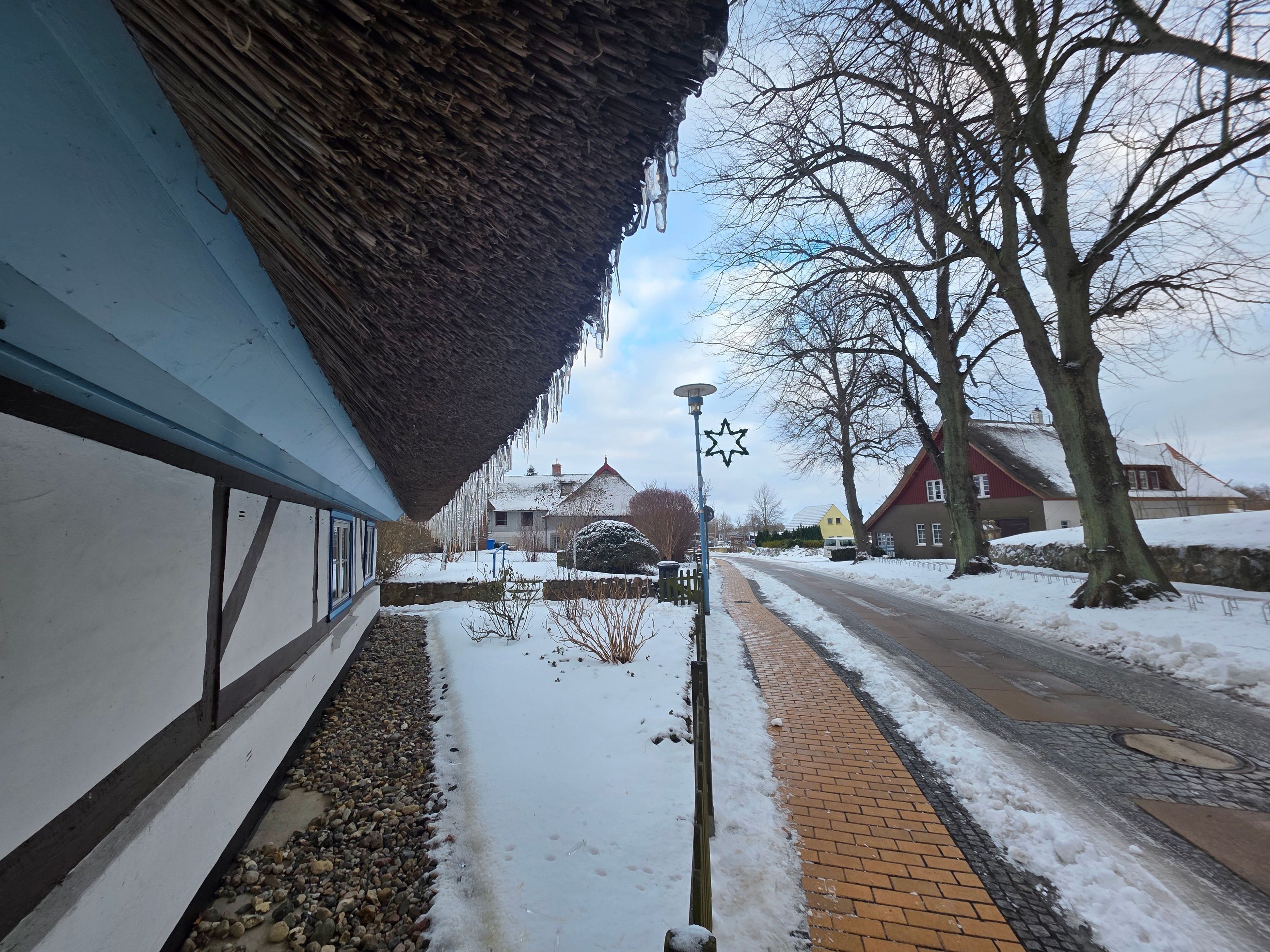 Une rue enneigée, bordée d’immeubles, avec un toit de chaume et des arbres sans feuilles.