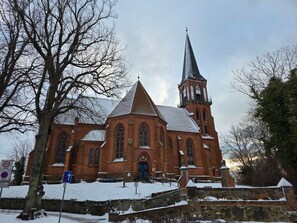 Une église en briques, dotée d’un clocher imposant et d’une tour horlogère, entourée d’un paysage enneigé et d’arbres sans feuilles.