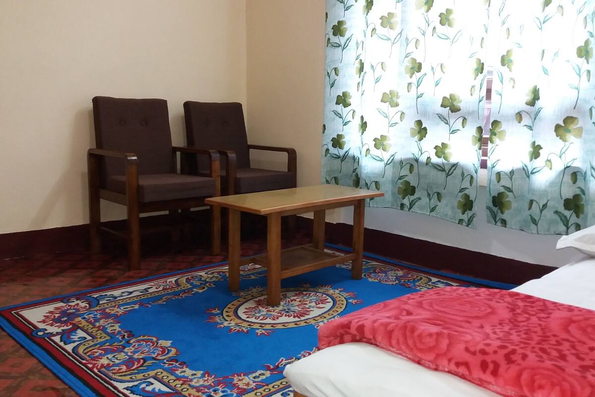 A room with two wooden chairs, a small wooden table, a patterned rug, a bed with a red blanket, and a window with floral curtains.
