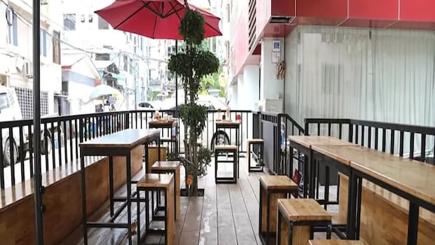 Un café al aire libre con mesas y bancos de madera, sombrillas rojas y vista a la calle.
