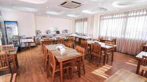 Un comedor amplio con mesas y sillas de madera, una cocina al fondo y ventanas grandes con cortinas traslúcidas.