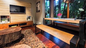 Een moderne woonkamer met een houten tv-meubel, een bank, een klein tafeltje met een bloevas en een groot raam met uitzicht op groen.