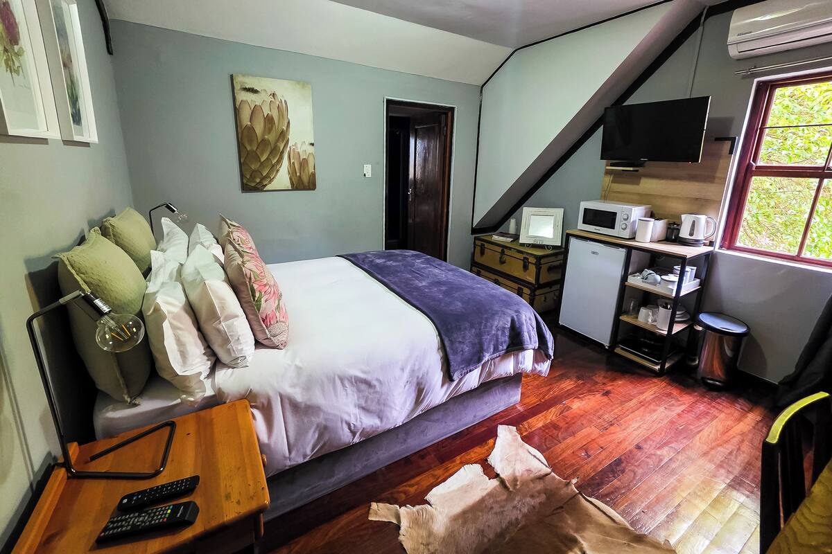 Un dormitorio con cama, almohadas, un escritorio con computadora, televisión y ventana.