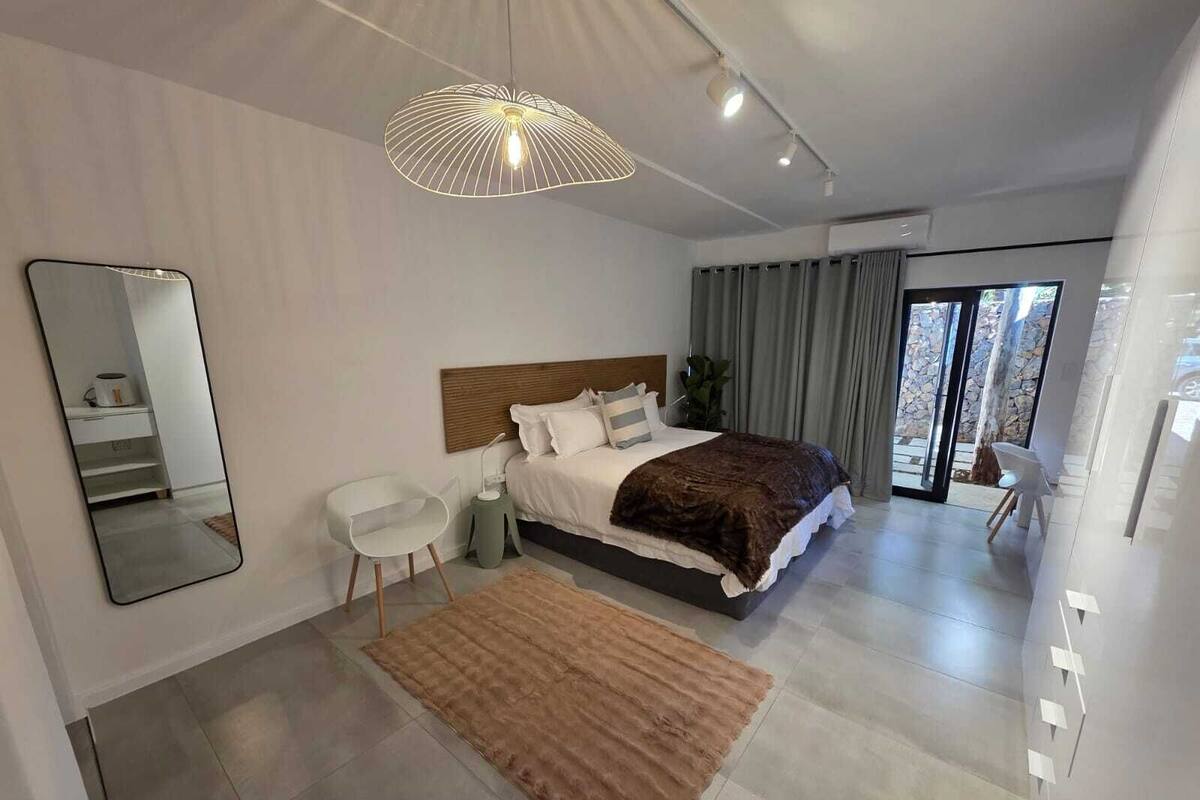 Un dormitorio moderno con una cama grande, una mesita de noche, una silla, un espejo y vistas al exterior.