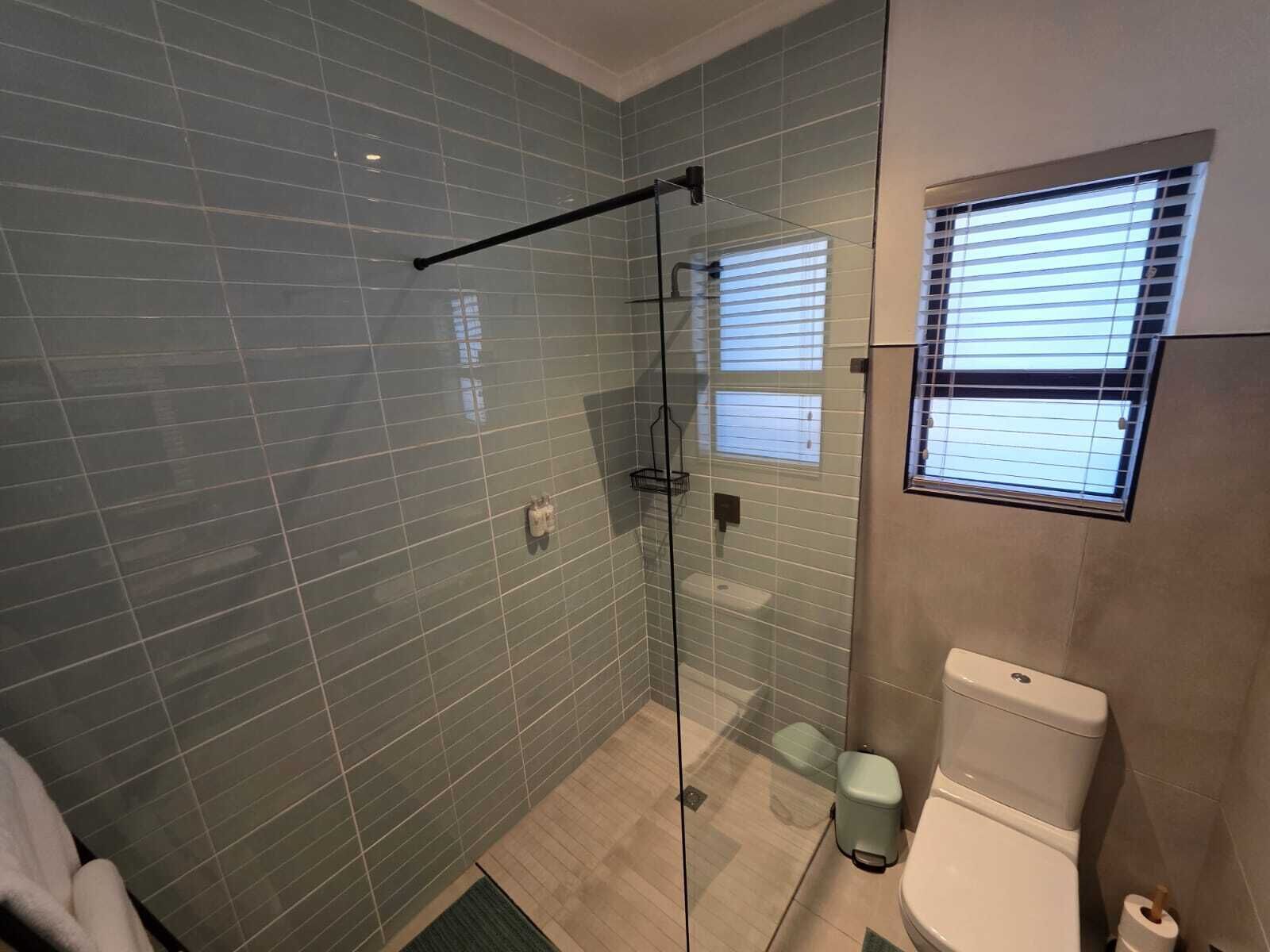 A modern bathroom with a glass shower enclosure, a toilet, and a window with blinds.