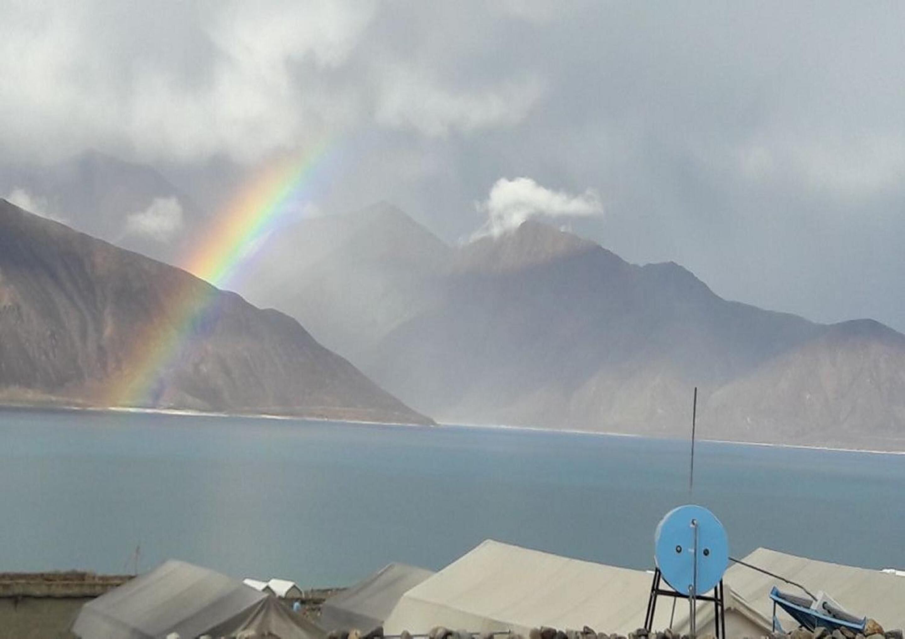 A satellite dish, tents, and a mountain range with a rainbow over a body of water.