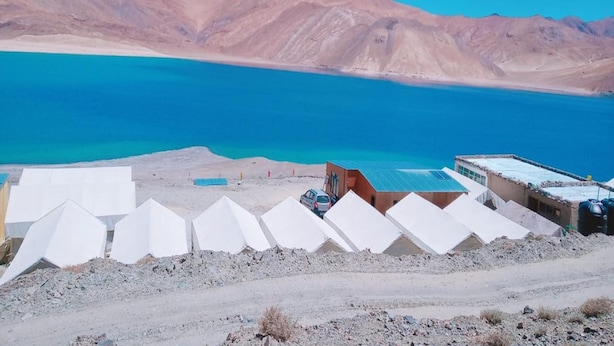 A desert landscape with a large body of water and mountains in the background. There are several white tents and a few buildings with flat roofs. - Eco Travellers Camp (Leh)
