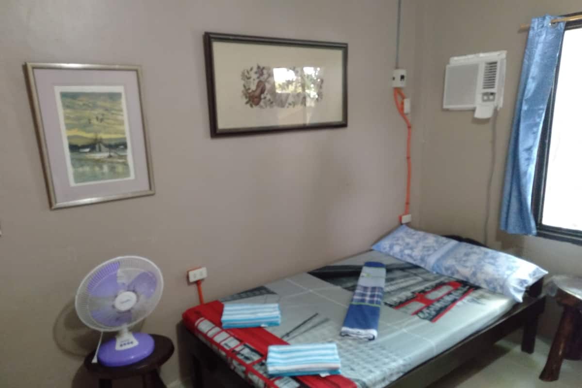 A small room with a single bed, a standing fan, a framed picture, an air conditioning unit, and a window with a curtain.