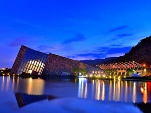 A modern building with a glass facade reflecting on a calm body of water, surrounded by a landscaped area with lights. - noir B&B (Toucheng)
