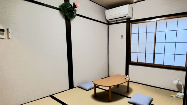 A tatami mat room with a low table, cushions, a hanging flower, an air conditioning unit, and a window with a grid pattern. - Hana Botan (Kyoto)