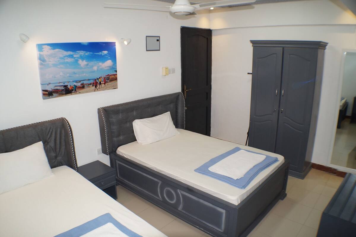 A bedroom with two beds, a wardrobe, a ceiling fan, and a beach scene painting on the wall.