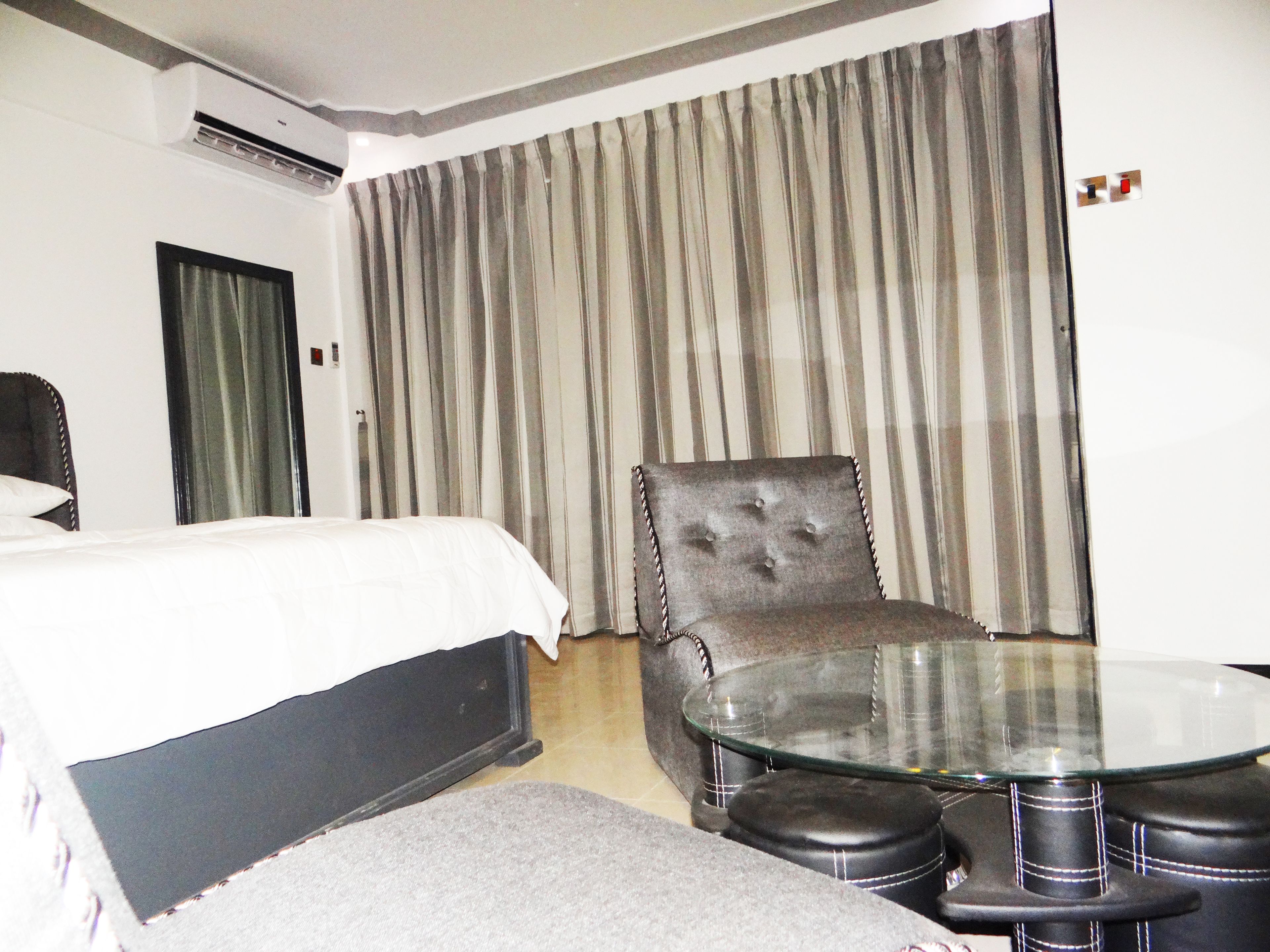 A hotel room with a bed, a tufted armchair, a glass-top table, and a small round table with stools.