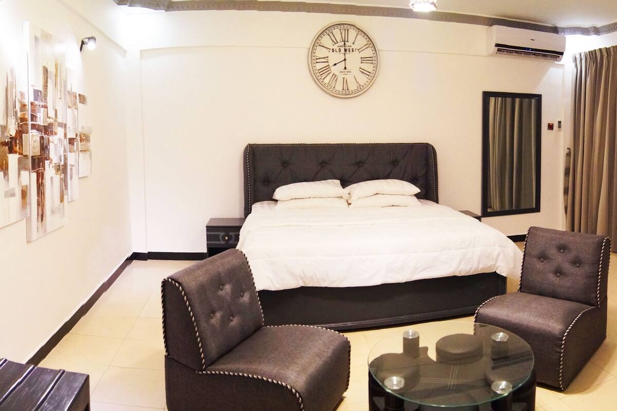 A bedroom with a large bed, two armchairs, a glass coffee table, a wall clock, and a curtain.