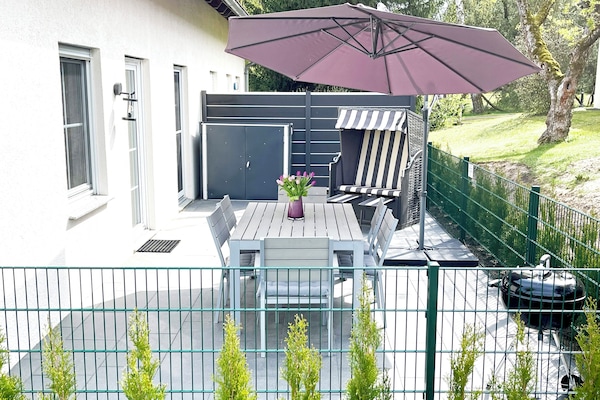 Umzäunte Terrasse mit Grill, Strandkorb, 2 Sonnenliegen, Esstisch und 6 Stühlen.