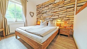 A bedroom with a stone wall, a bed with checkered bedding, a bedside table with a lamp, and a window with curtains. - Fantastically situated vacation home for families & dogs, fenced, 200m to Lake Plau (Fünfseen OT Lenz-Süd)