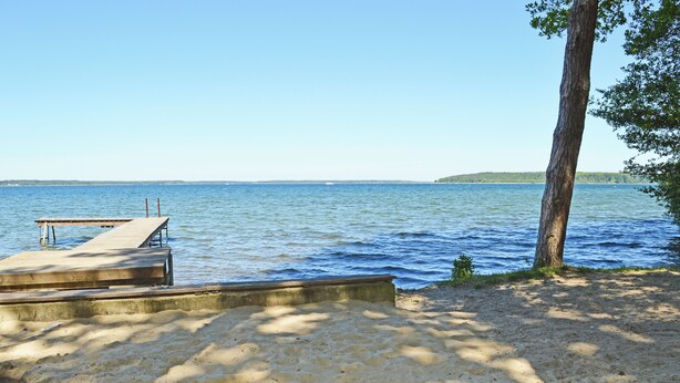 A sandy beach with a wooden dock extending into calm water, surrounded by trees. - Fantastically situated vacation home for families & dogs, fenced, 200m to Lake Plau (Fünfseen OT Lenz-Süd)