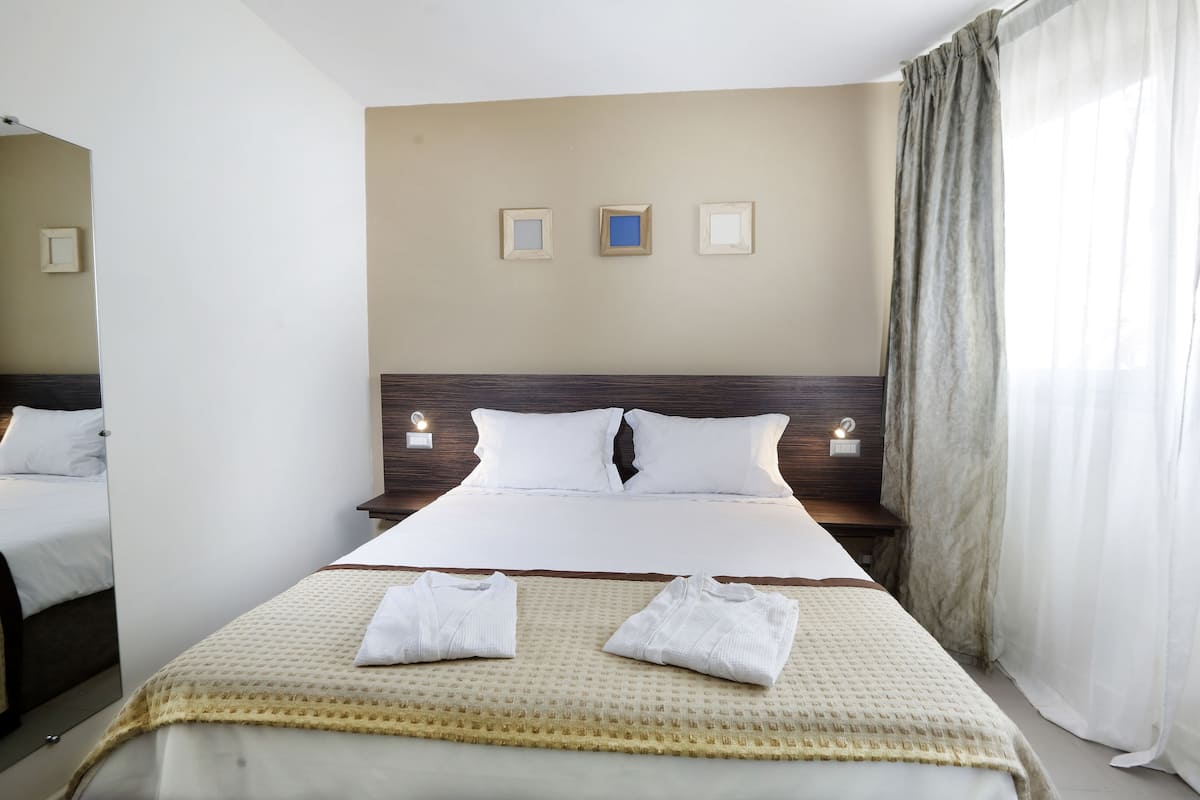A neatly made bed with white linens, a beige throw blanket, and two folded white towels. There are three wall-mounted light fixtures and a large window with sheer curtains.