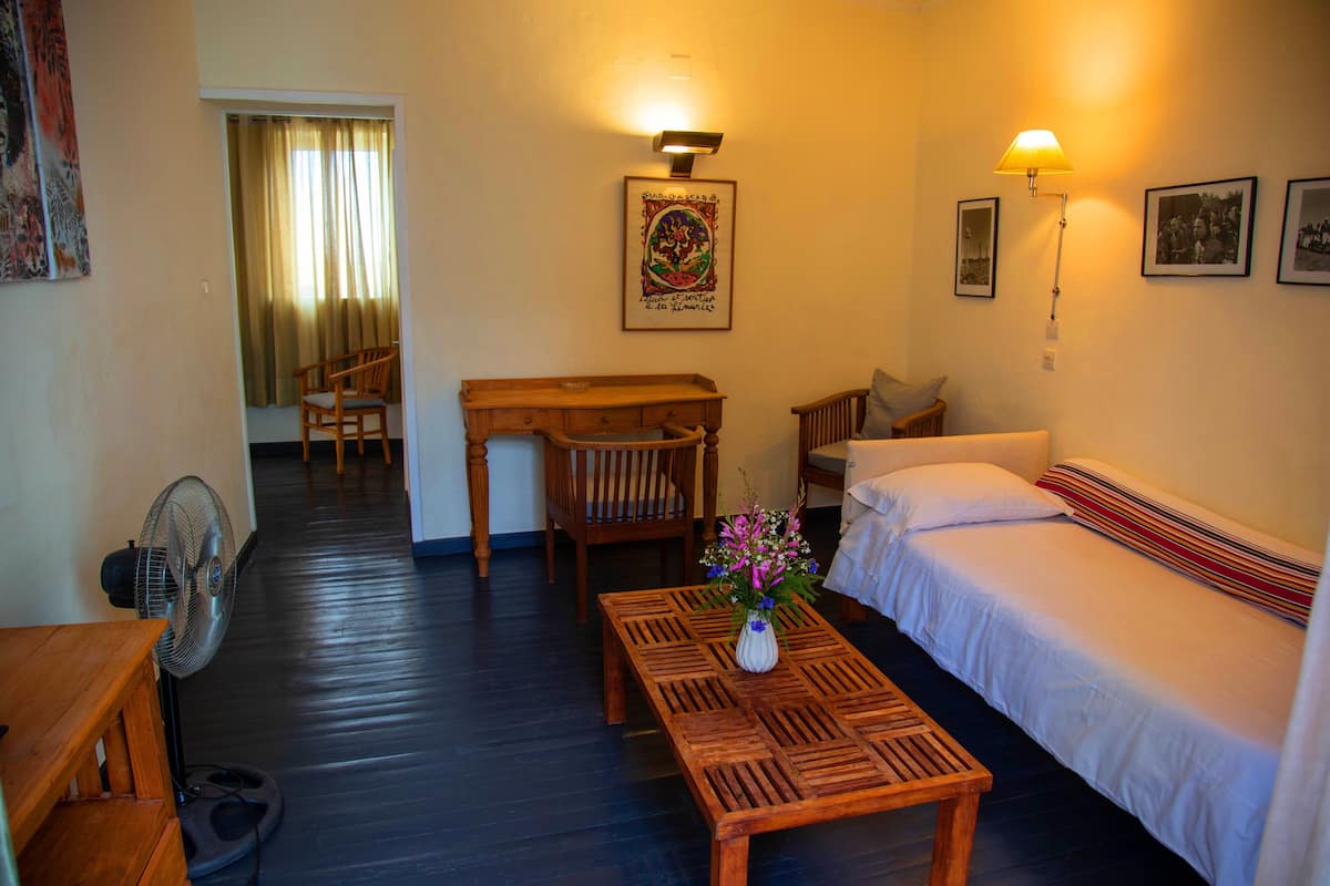 A hotel room with a bed, a wooden table with a vase of flowers, a chair, a desk, a fan, and a lamp.