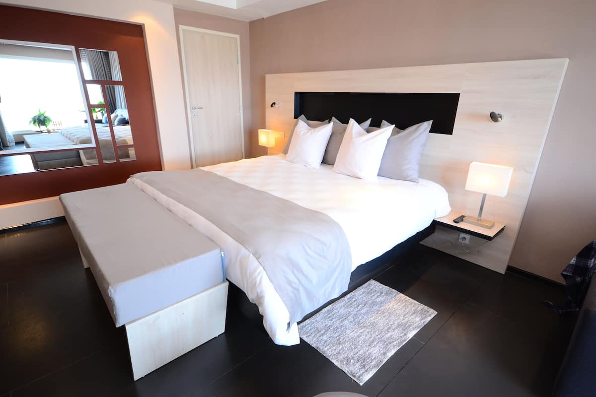 A modern hotel room with a large bed, bedside tables with lamps, a bench, and a view through a window.