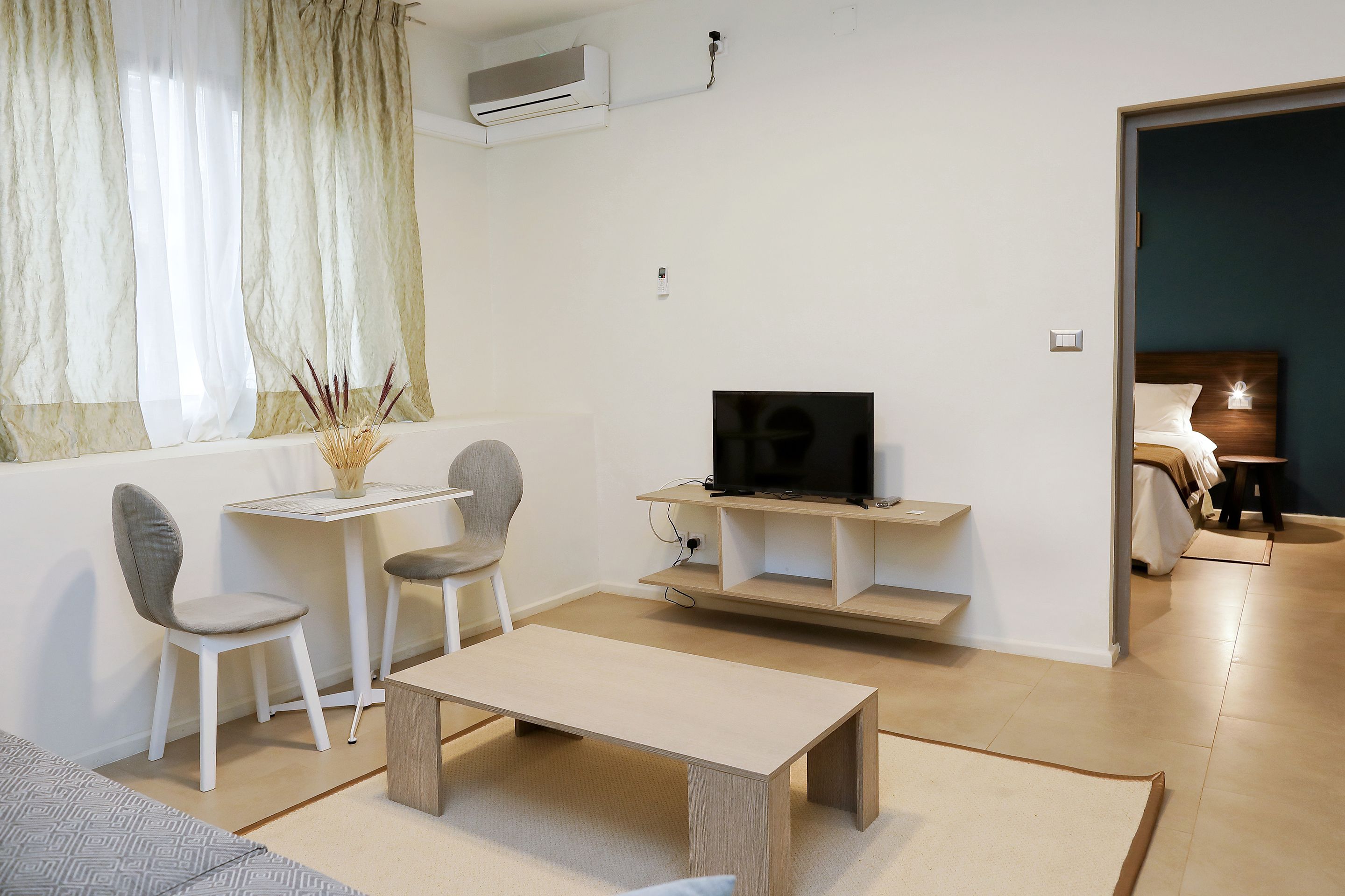 A modern living room with a TV, a coffee table, two chairs, a small table, and a bed in the adjacent room.