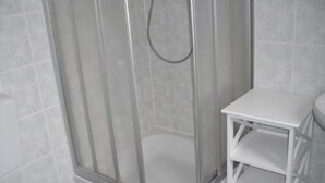 A shower enclosure with a glass door, a white tiled wall, and a white shelf unit beside it. - Vacation apartment MAXIM - Holiday apartments (Koserow)