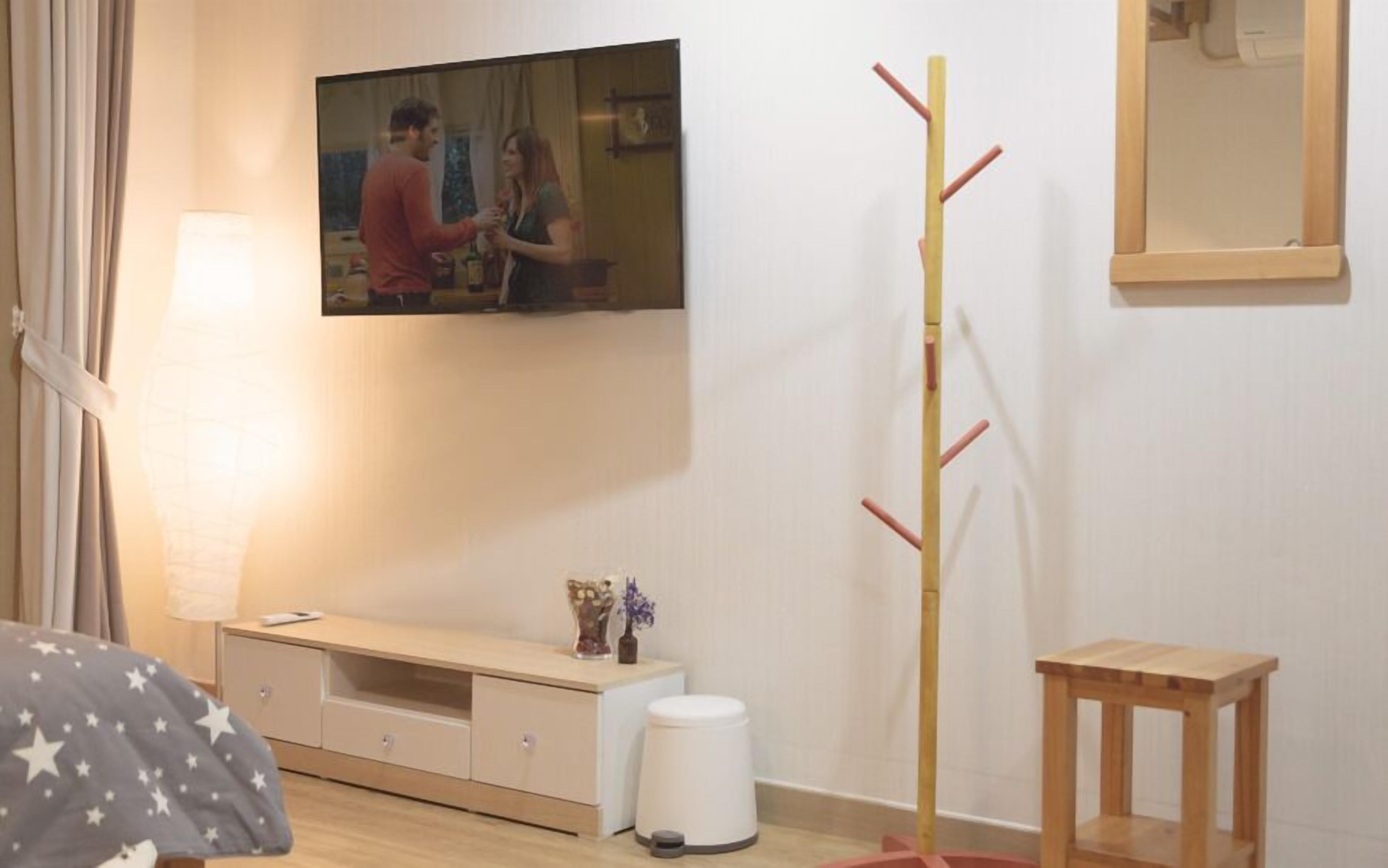 A room with a TV mounted on the wall, a wooden coat rack, a small wooden table, a bedside table with drawers, and a lamp.