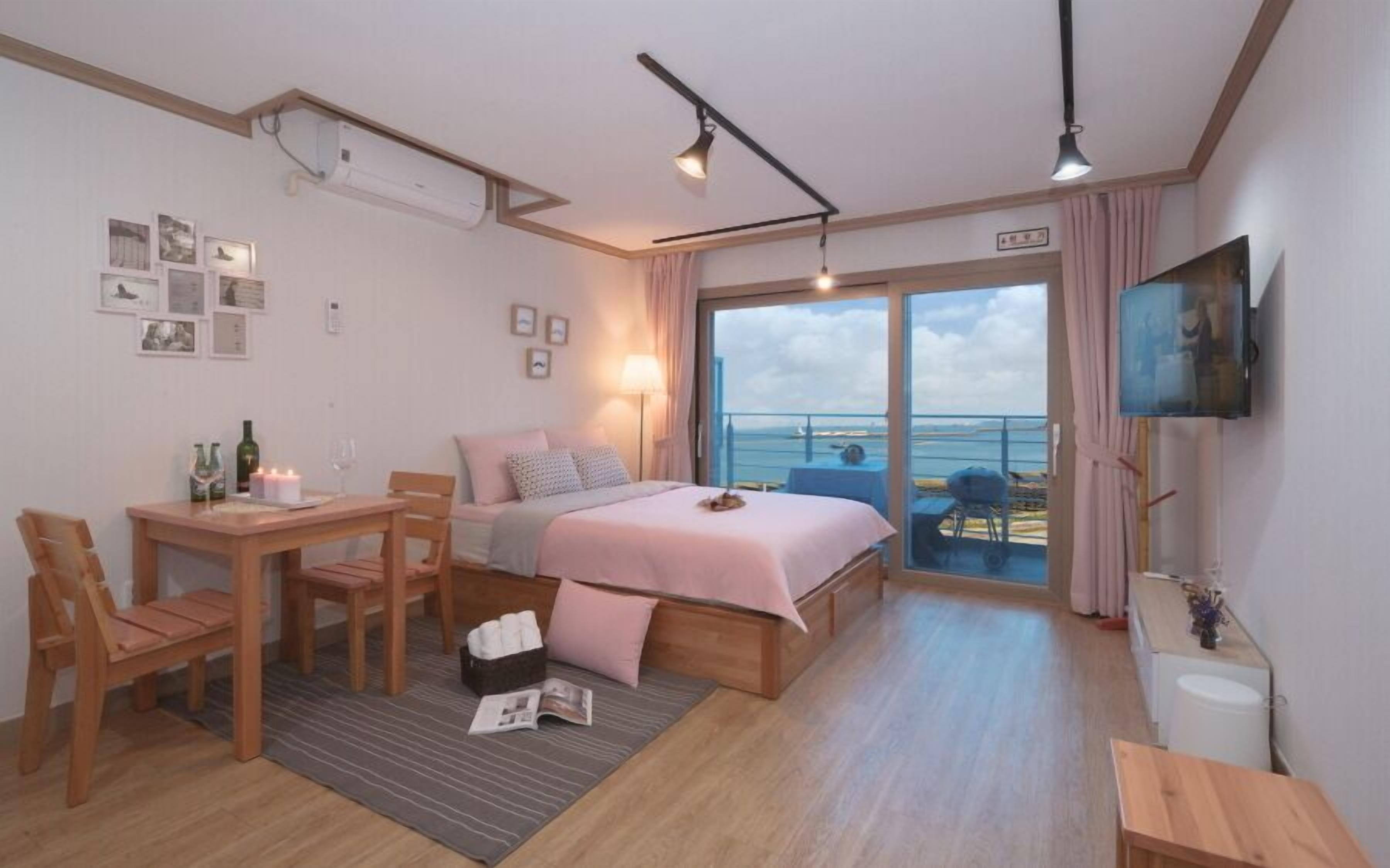 A hotel room with a bed, a desk, a chair, a TV, and a balcony with a view of the sea.