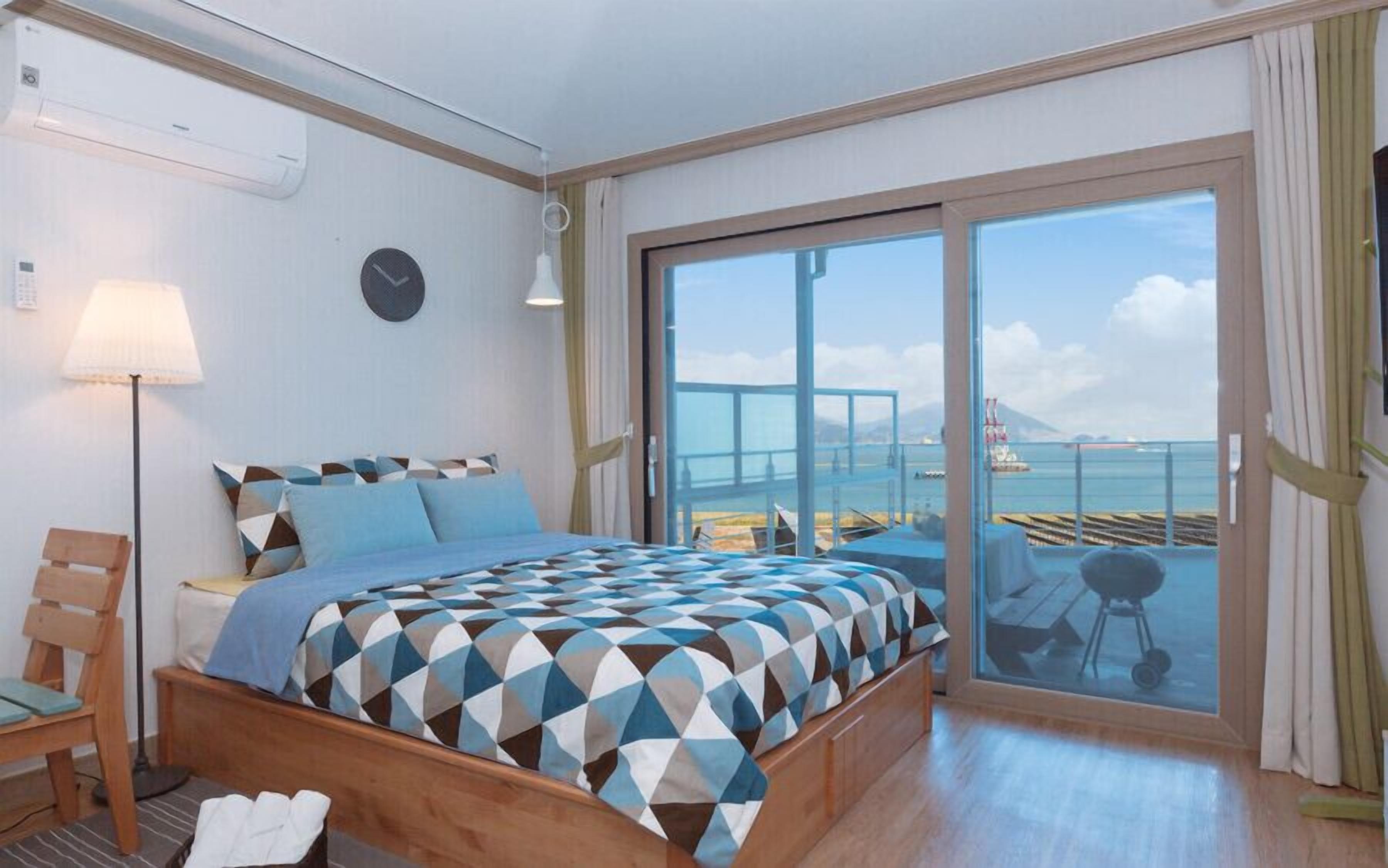 Habitación de hotel con cama, una silla, una lámpara, un reloj y un amplio ventanal corredero que da a un balcón con vistas al mar y una montaña distante.