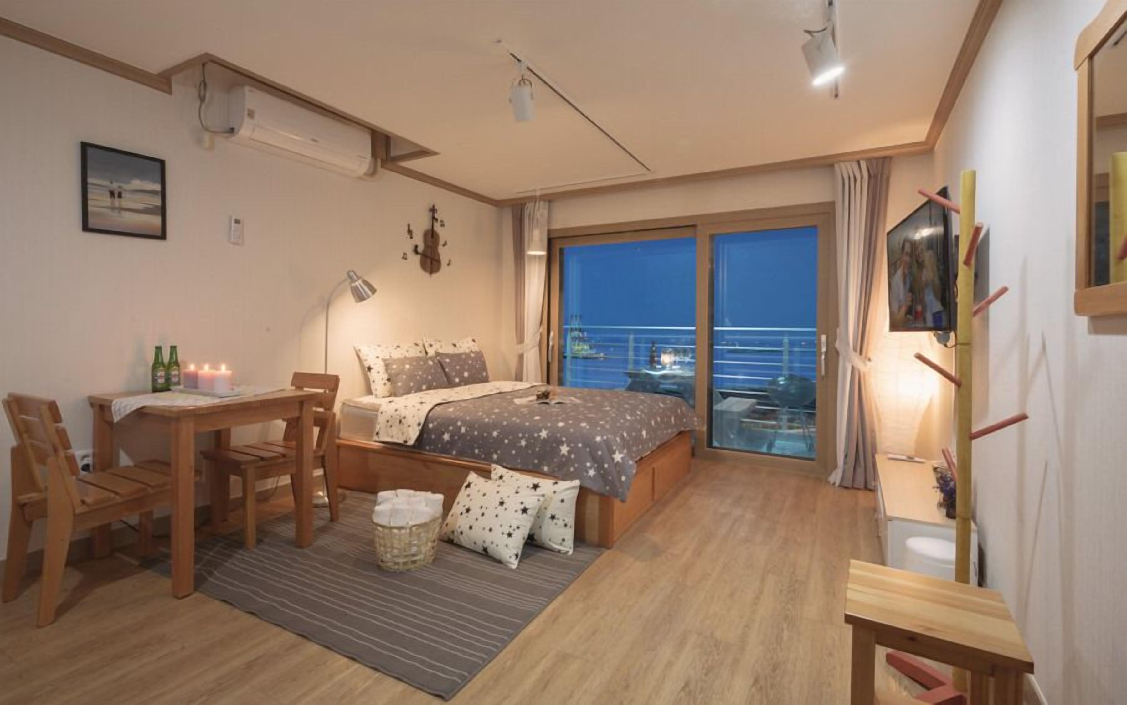 A cozy bedroom with a bed, bedside table, and a desk with a chair. There is a window with a view of the sea and a balcony.