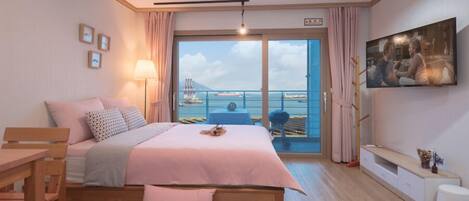 A bedroom with a bed, a desk, a chair, a TV, and a balcony with a view of the sea.