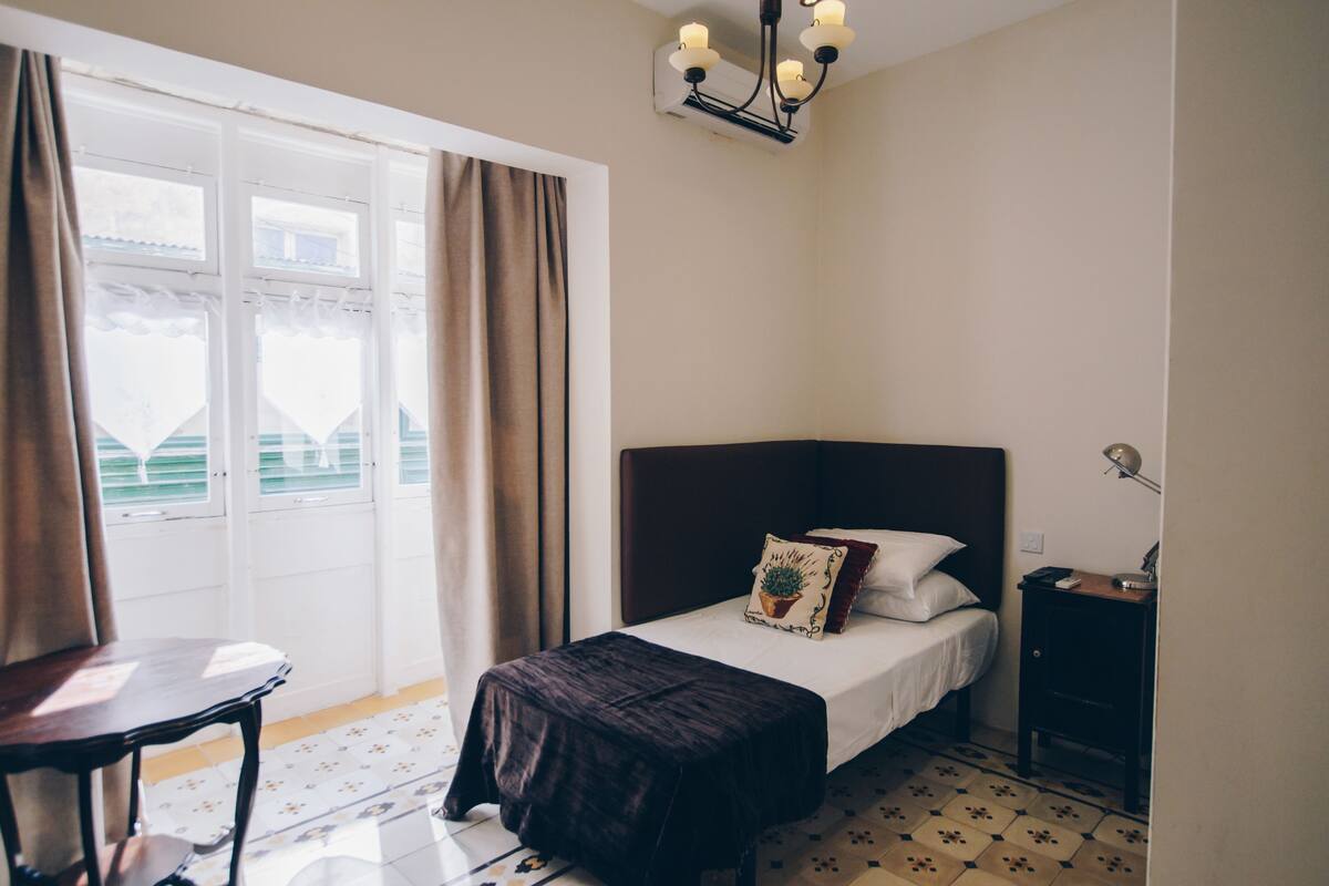 A bedroom with a bed, a nightstand, a table, and a window with curtains.