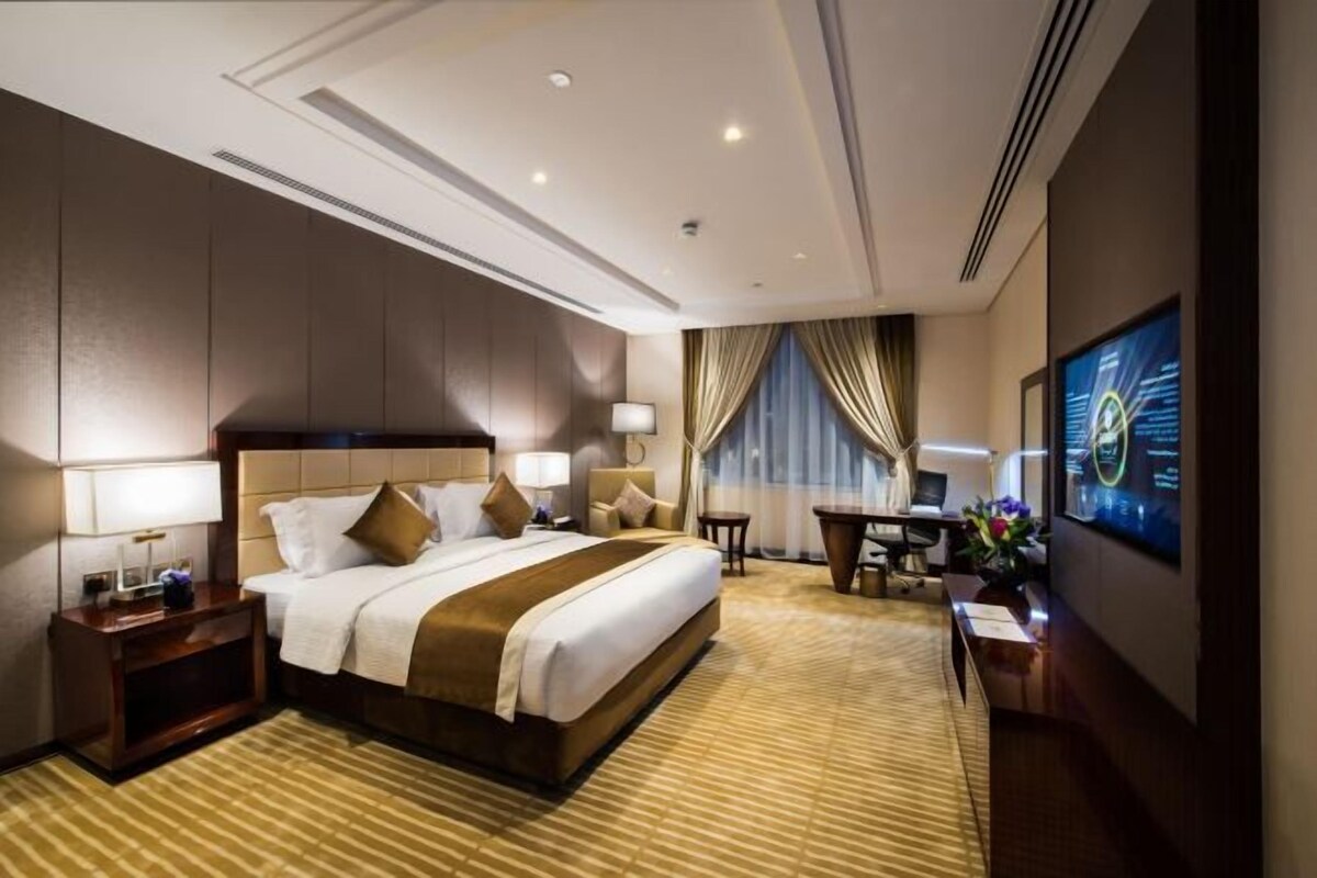 A hotel room with a large bed, a desk, a chair, a TV, and a desk lamp.