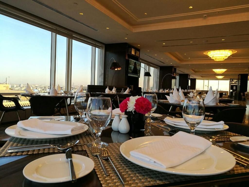 Un comedor con mesas dispuestas para dos, ventanas amplias que ofrecen vistas a la ciudad y una araña colgando del techo.