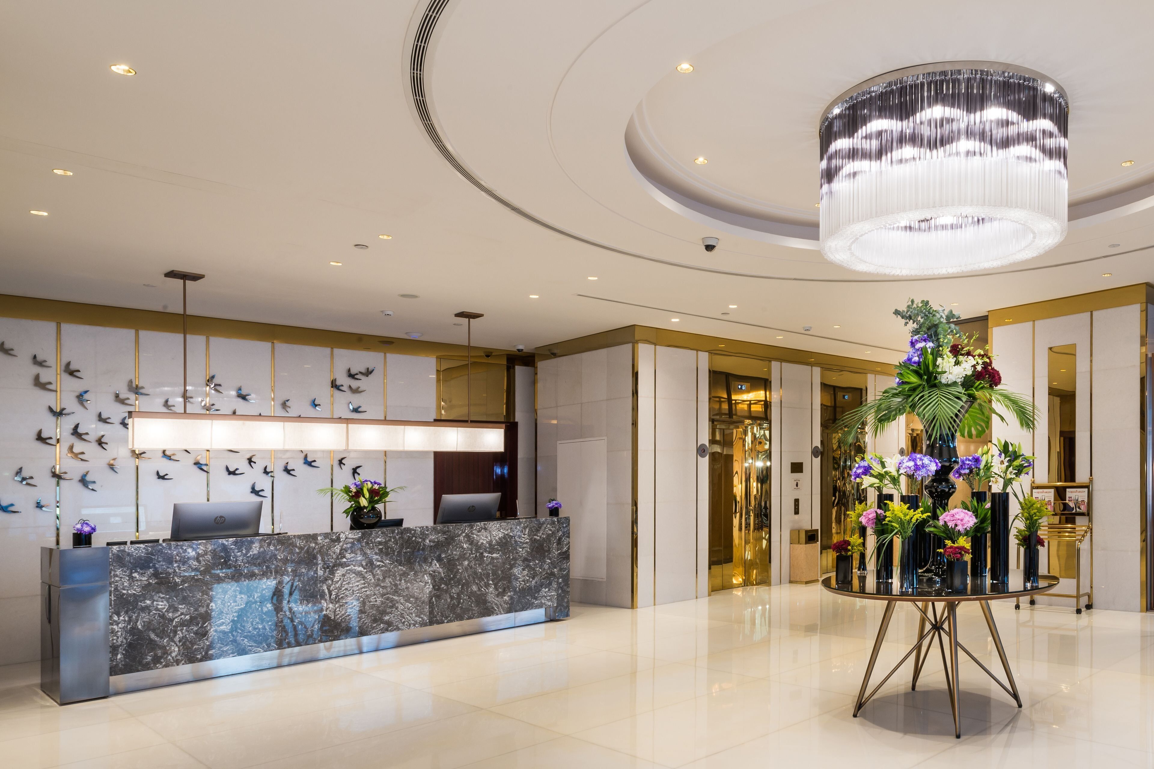 A modern hotel reception area with a marble counter, a large chandelier, and a wall-mounted bird display.