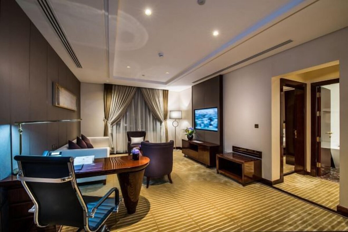Ein modernes Hotelzimmer mit einer Couch, einem Sessel und einem kleinen Tisch. Ein Fernseher ist an der Wand montiert und eine Tür führt in einen weiteren Raum.